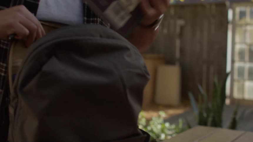 A teenager in a plaid shirt and white tshirt putting notebooks into his backpack and getting off the table