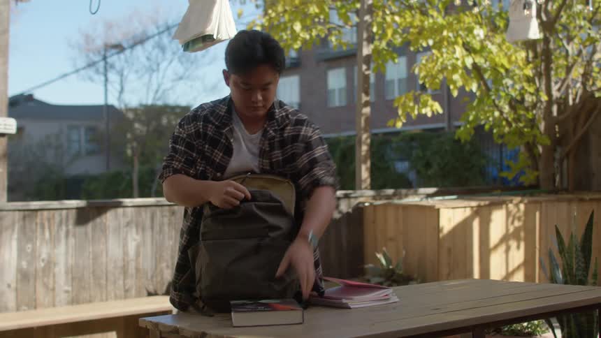 A young man sitting at an outdoor bench stands up and packs his backpack