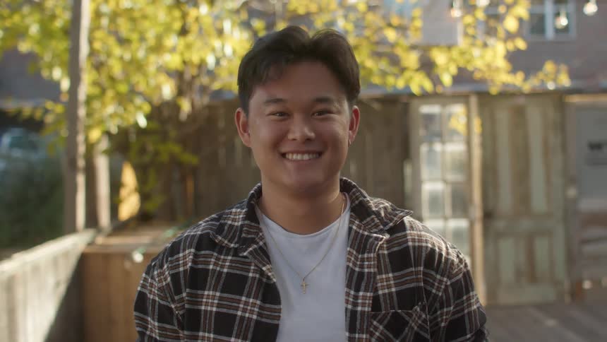 Man wearing a plaid shirt white t shirt and a necklace smiles at camera in the yard