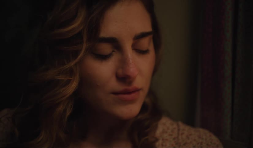 A sad woman with brown curly hair wearing a white shirt gazes downward crying