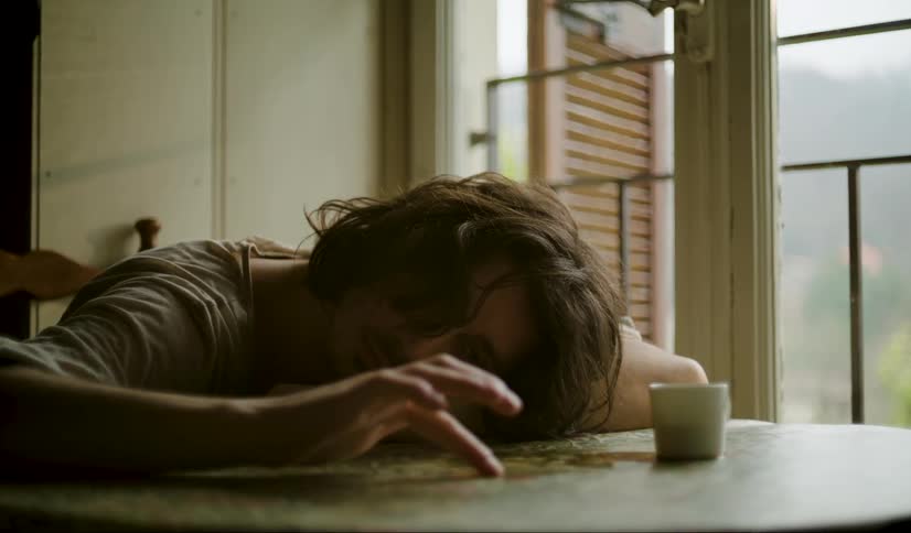 Man leans his head and arms onto table and plays with spilled coffee on a foggy day