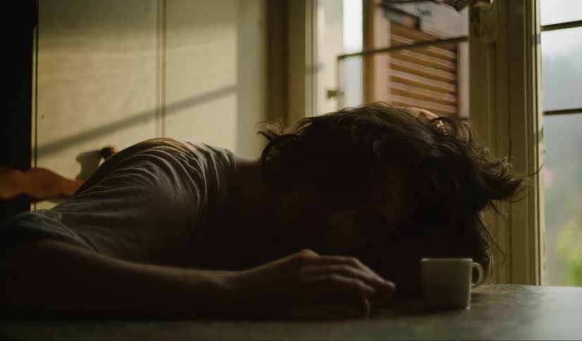 A tired man with long hair wearing a white tshirt sits at a table