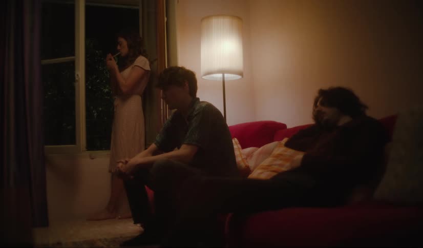 A woman smokes by the window while two men sit on a couch looking bored