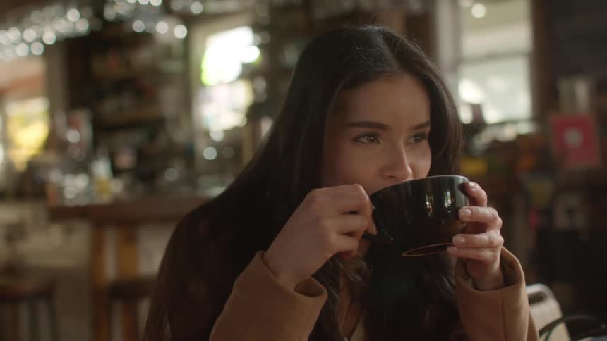 A woman in a coffee shop seated smiling while sipping a cup of coffee