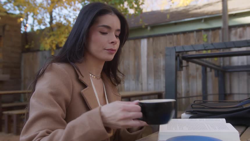 A woman is sitting outside at a table drinking a cup of coffee and reading a book