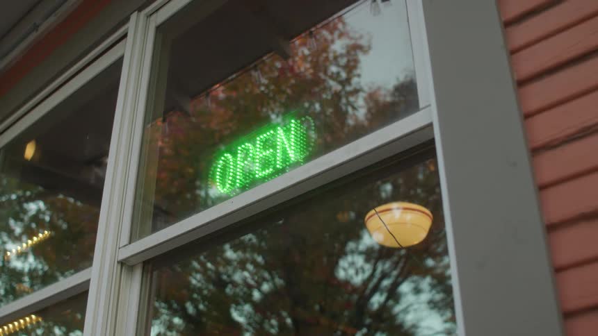 Hand reaches up and switches a neon sign in a restaurant store front window from red letters that read closed to green letters that read open on a sunny fall day