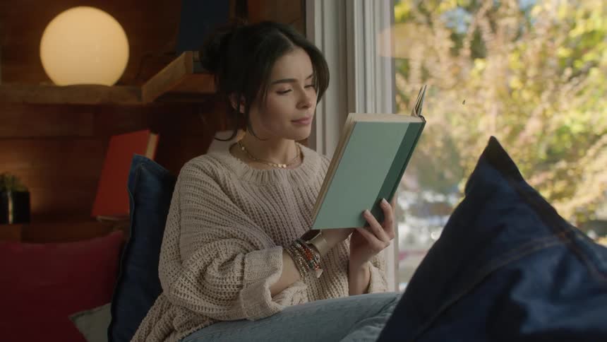 A woman reads a book while sitting next to a large window