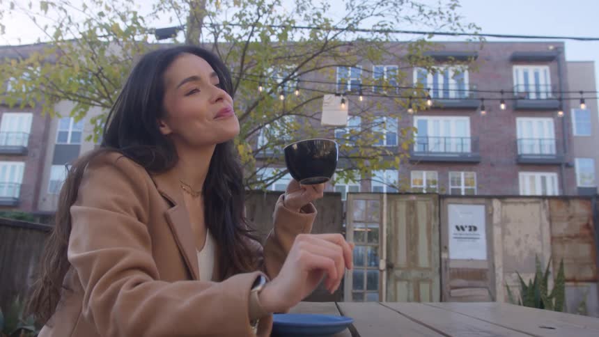 A woman with straight hair and a brown jacket sitting at a table in a cafe yard casually sipping from her cup