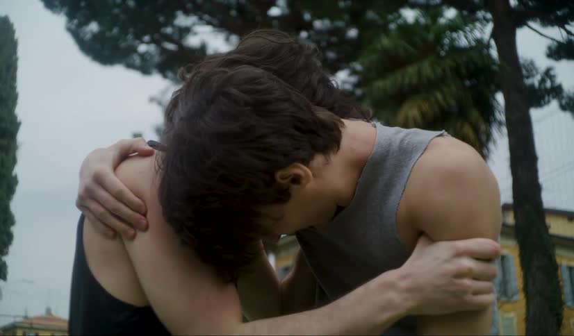 Two men embrace each other while slow dancing on a court outside some yellow buildings