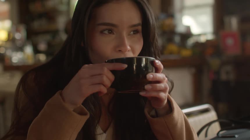 Woman with dark hair a brown coat and a white shirt sips from black cup sitting at a table in the cafe