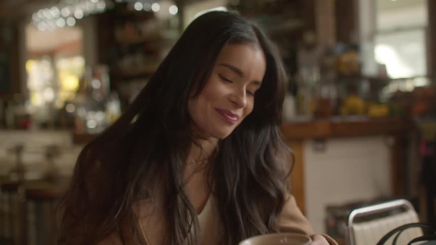 Brunette woman sits at a table in a coffee shop and makes a sour face after she takes a sip from a large brown cup followed by talking setting the cup on a saucer on the table and pushing back her bangs