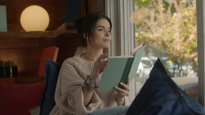 Woman with a white sweater reads a book by the window