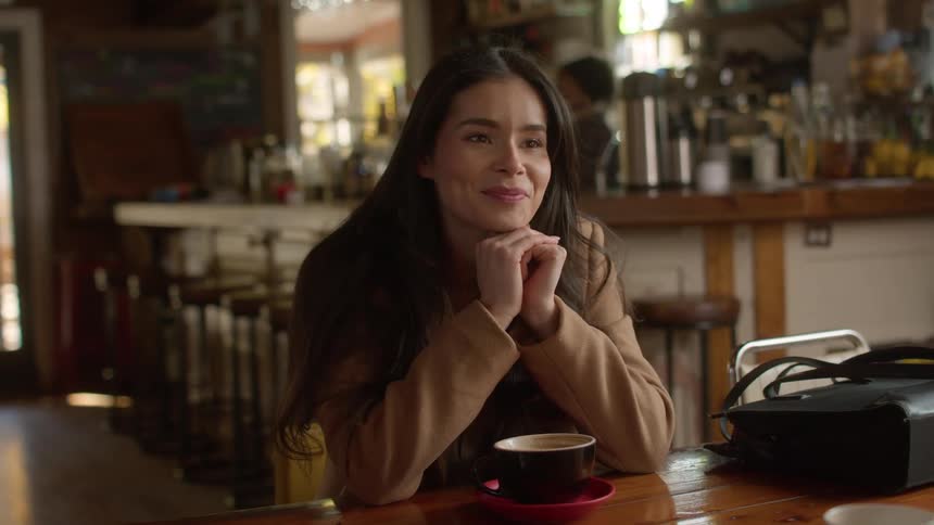 A woman sitting at a coffee shop table opposite another smiles and tilts her head as she talks and listens