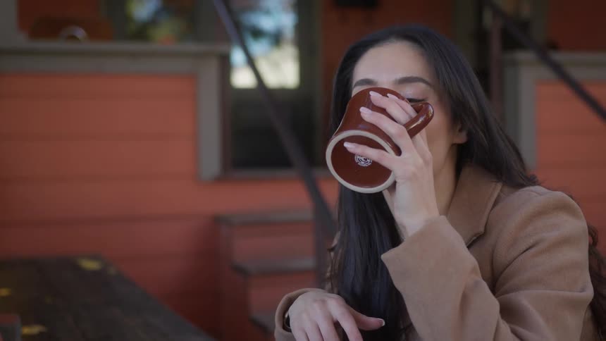 A woman with straight hair sporting a brown coat sits at a table calmly sipping coffee from a brown cup in the cafe