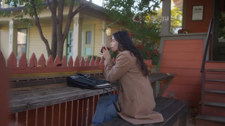 A woman is sitting on a bench in her backyard and is drinking something