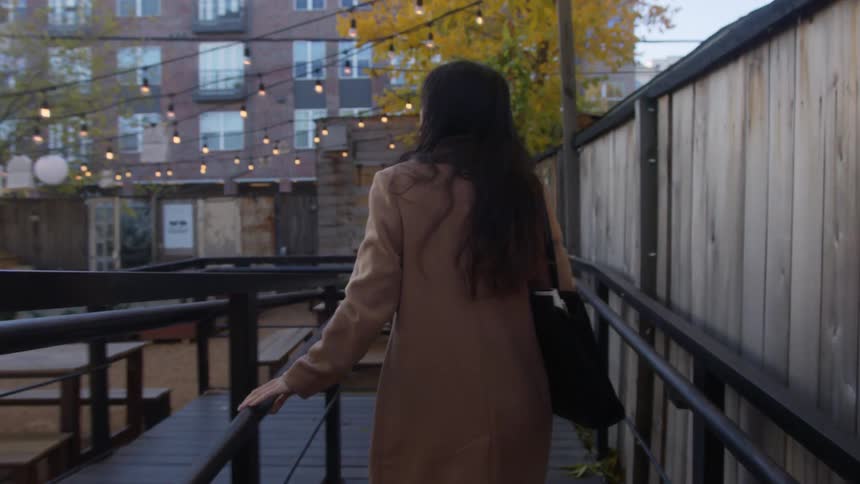 A young woman wearing a trench coat is walking forward and leaning on a wooden fence