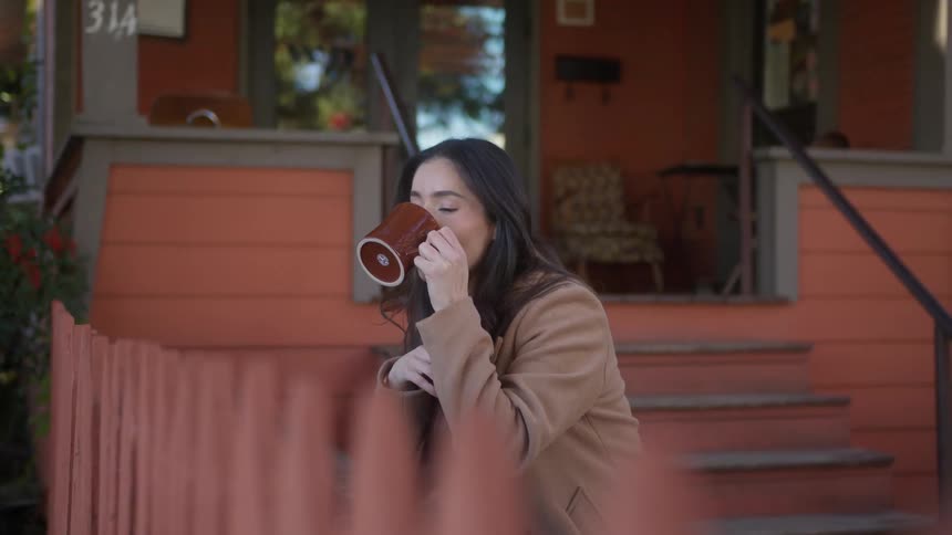 Lady sits outside of a house at midday drinking from a mug