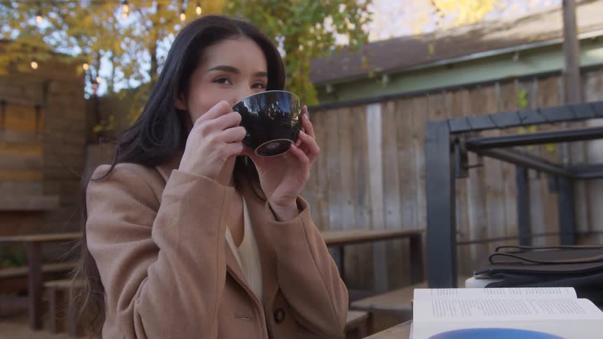 Brunette woman sits outdoors sips from mug book on table