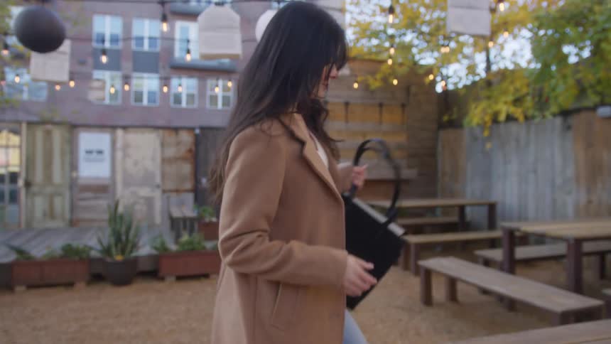 Happy woman walks through the outdoor dining area and sits at a table as she places her purse on top of it