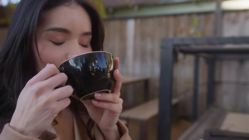 A woman with dark hair takes a sip from her coffee while seated outdoors