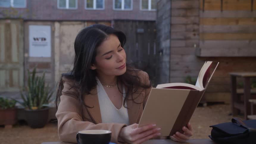 Woman sits at an outdoor table with a book and a mug