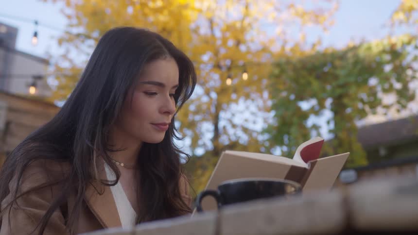 Lady reads a book while seated outdoor at a table with a mug during the day