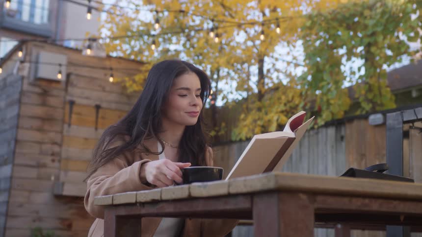 A woman drinks coffee while reading a book outdoors