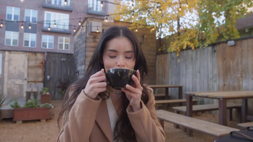 Woman with long brown hair at a table in a restaurant backyard outdoor seating area closes a book picks up a coffee cup with both hands takes a sip then looks around with a content smile on a fall day in the late afternnon