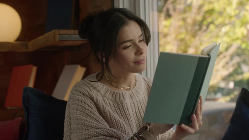 Woman wearing a white sweater and jeans reads a book by the windows