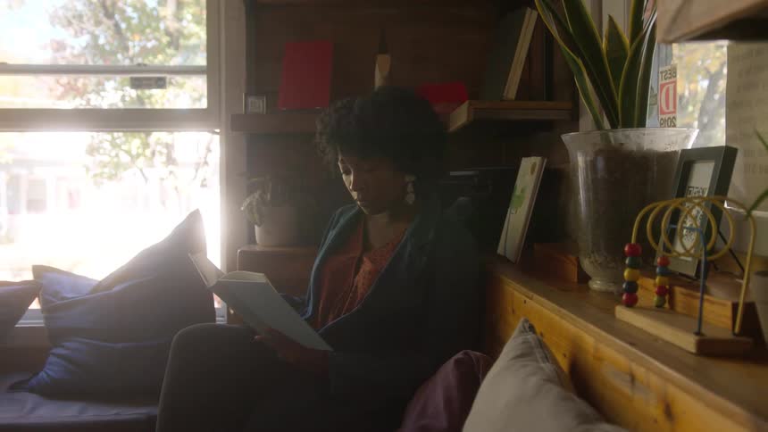 A young woman sits indoors near an open window while reading