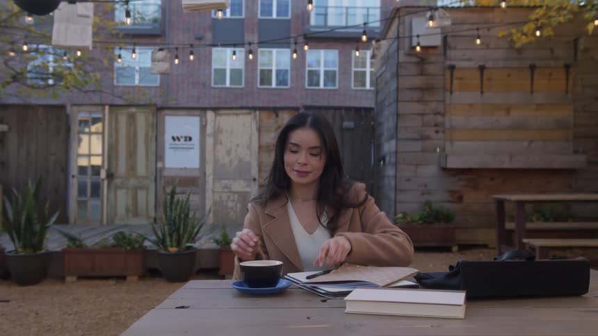 A woman with straight hair and a brown jacket sitting at a table in a cafe yard casually sipping from her cup