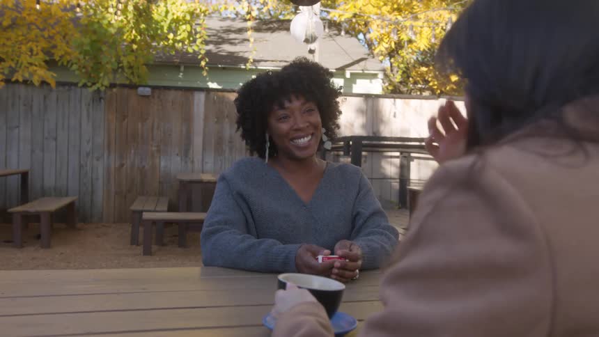 A woman carrying a deck of cards sits down on a bench opposite another in a restaurant outdoor seating area