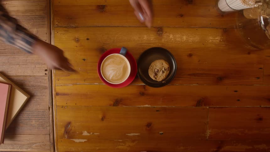 Hands placing a coffee and a cookie on a wooden table and then being picked up by a person