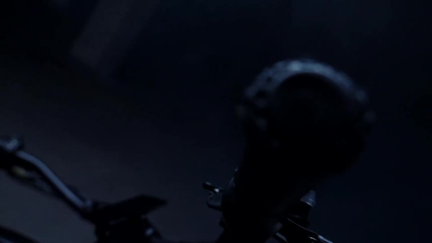 A black motorcycle sits in an all black room focusing on the handlebars