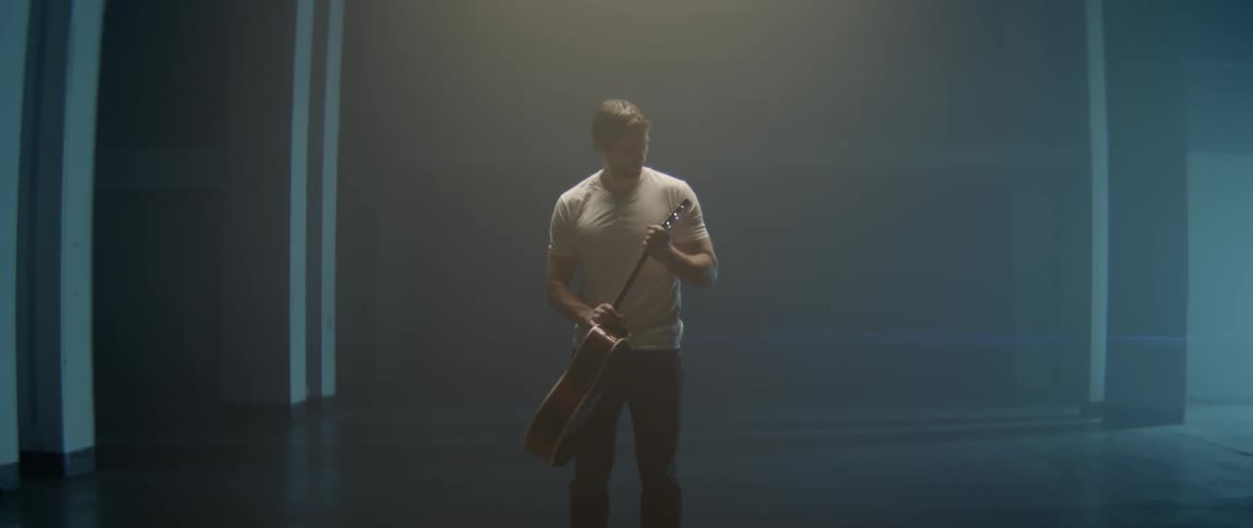 A man in a white t-shirt carefully examines an acoustic guitar in a studio.