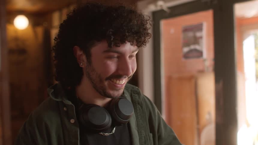 A man with curly hair a beard and a green jacket wearing headphones carries two cups of coffee to a table in a cafe