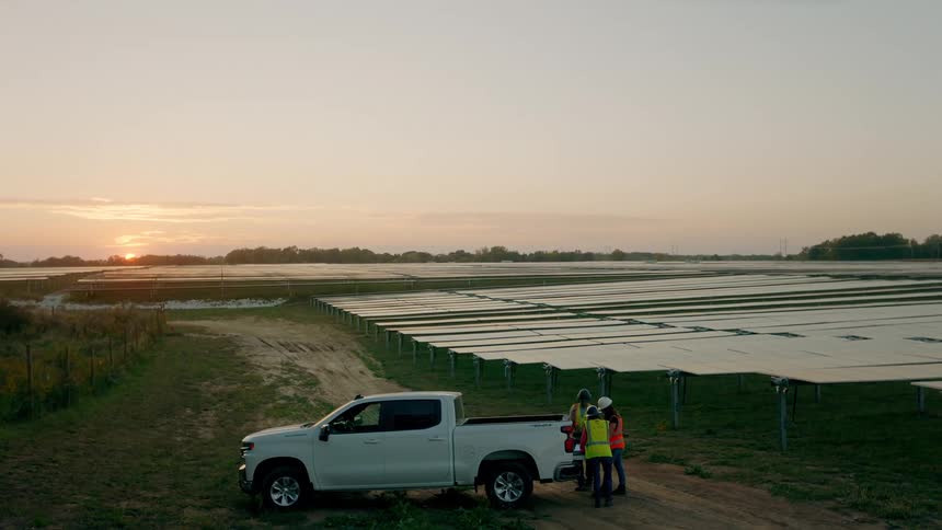 Three men wearing green and red vests stand behind a pickup truck at sunset