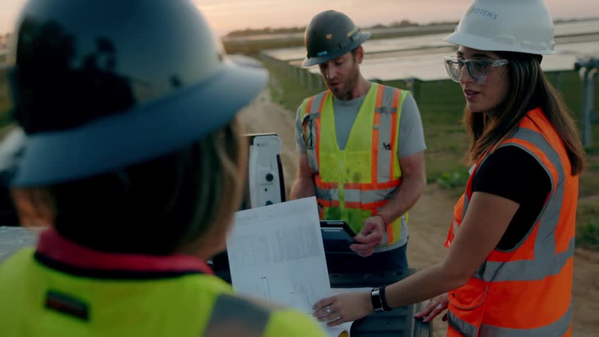 Coworkers consult plans lying on truck bed parked on dirt road at sunset among rows of solar panels
