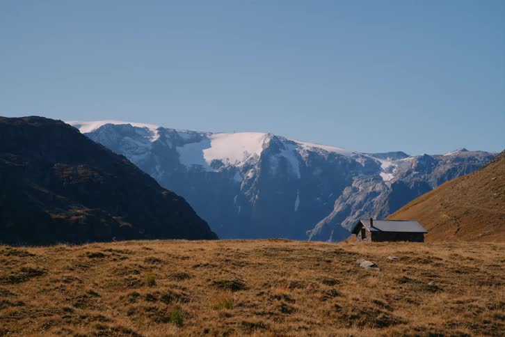 A wood cabin sits on the top of a mountain overlooking a snowcapped mountain