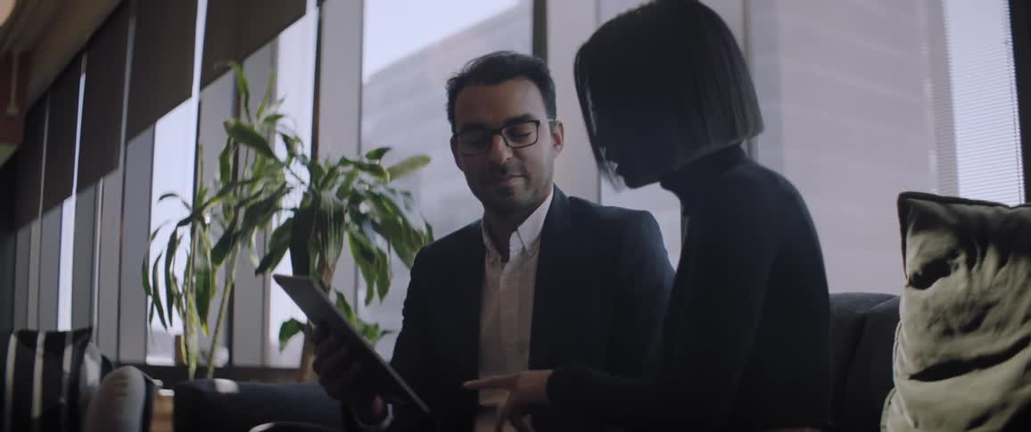 A man and a woman are talking and the woman points to the tablet in the mans hand