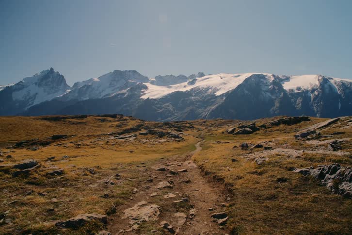Dirt trail through rocky moss covered highlands with snow covered mountain range and blue sky in the background on a bright day