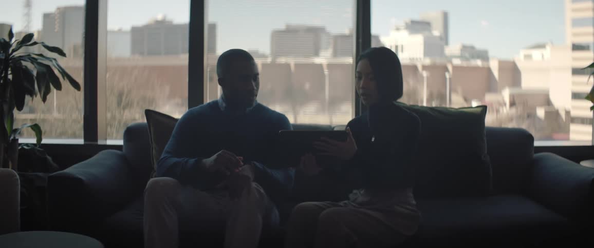 A man and a woman sitting on a sofa talking while the woman shows something on a tablet to the man