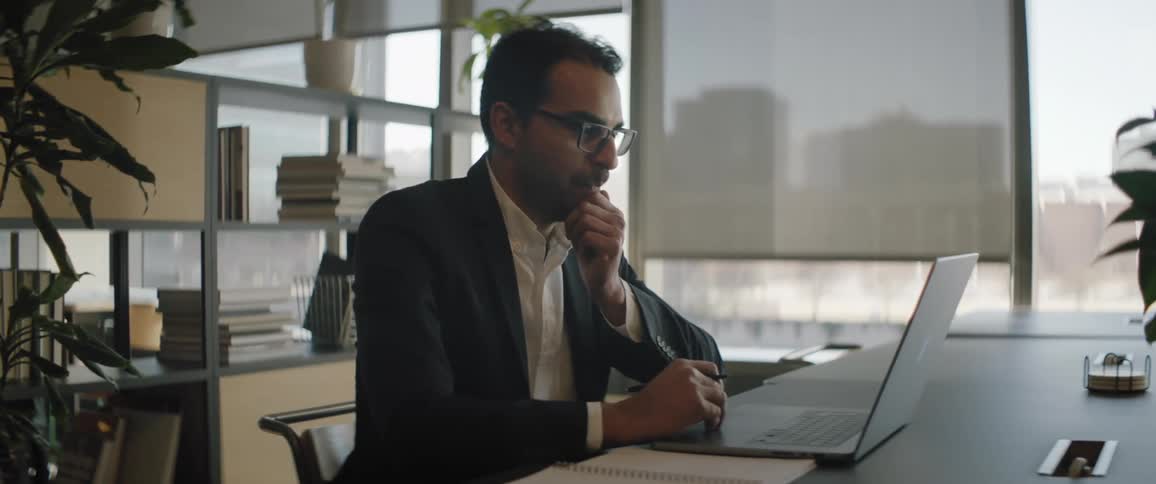 A man is sitting at a desk in an office working on a laptop computer as he stops to think and rubs his chin