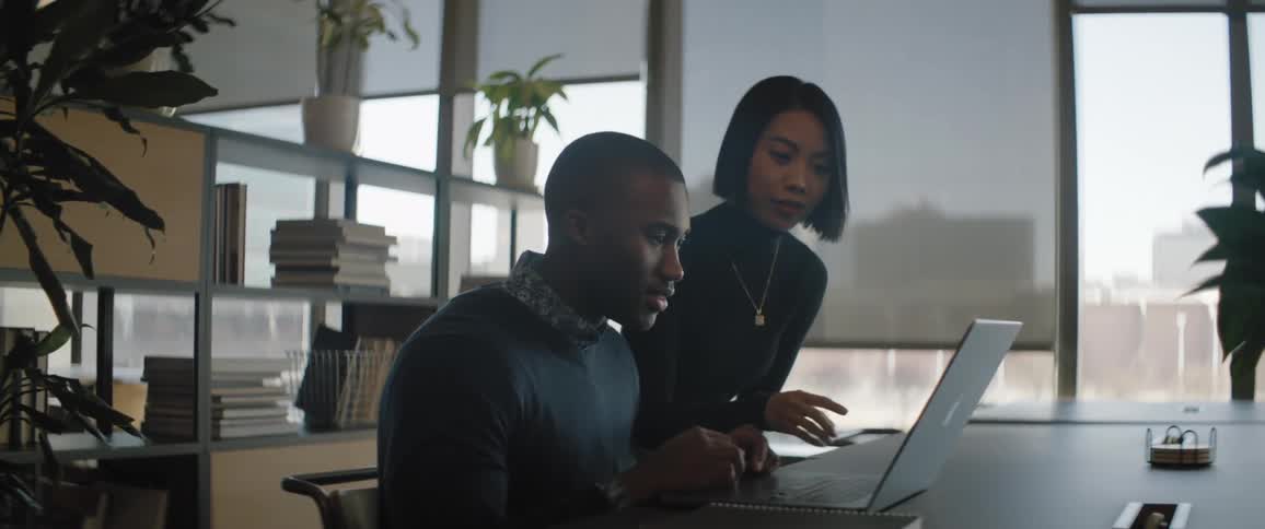 A man sits and uses a laptop while talking to a woman next to him