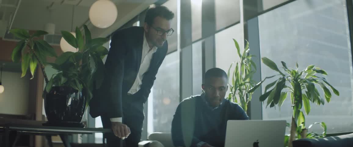 A man standing next to a man who is sitting and using a laptop on a table