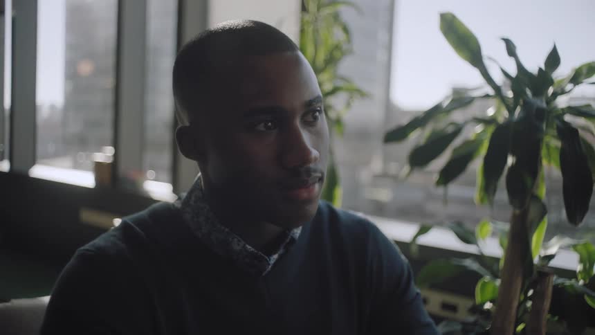 A black male with a polo sweater over a dress shirt sits expressionless in an office building beside a row of windows on a bright day