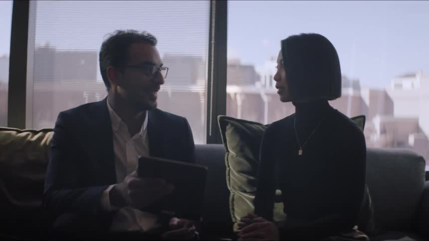 Male and female sit on a couch in a building downtown in front of large windows talking while the man holds a tablet and the city skyline is in the background