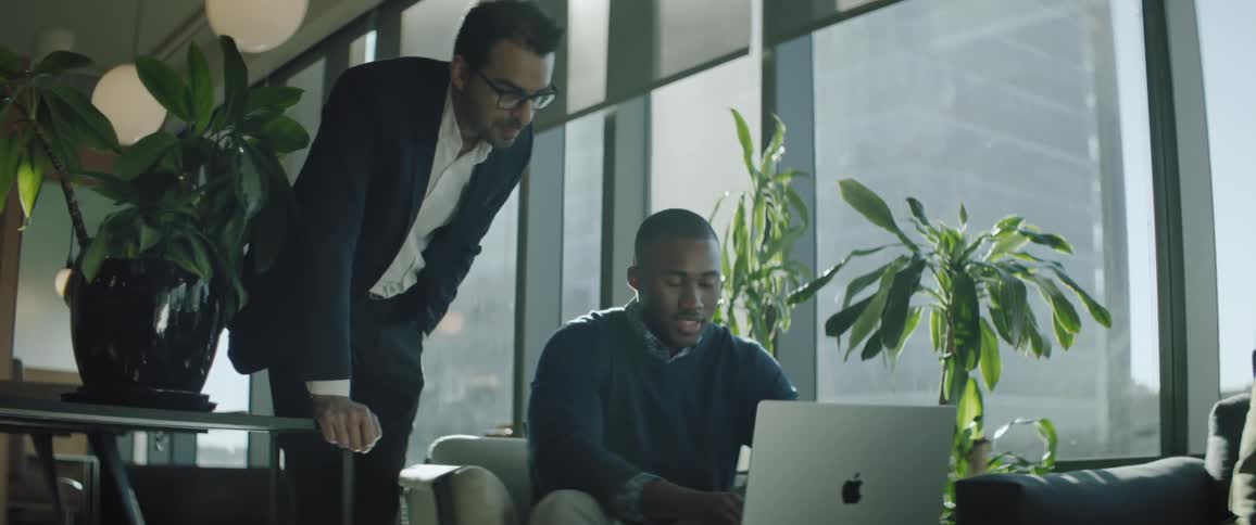 A black man seated in front of a laptop talking with another man standing behind him inside an office during the day