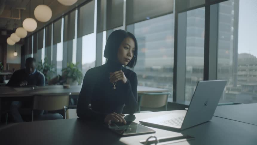 A seated woman is using a tablet and a laptop that are on a table