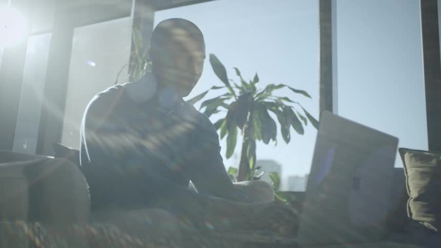A man typing on a laptop while seated on a chair indoor at midday